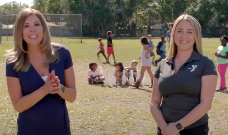 Still from video of Gayle Guyardo and Alyssa Heartstock outside in empty field, smiling. Kids playing games in after school program in the background.